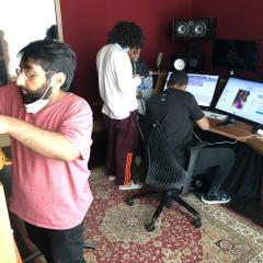 Candid photo of four people in a sound studio, in front of computer monitors displaying music editing software