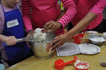 Two children scoop icing off of a whisk into a metal bowl, the instructor leans over the help