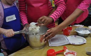 Two children scoop icing off of a whisk into a metal bowl, the instructor leans over the help