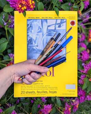 Photo of a hand holding a fistful of pens, pencils, and brushes in front of a pad of bristol paper. Flowers and leaves frame the image